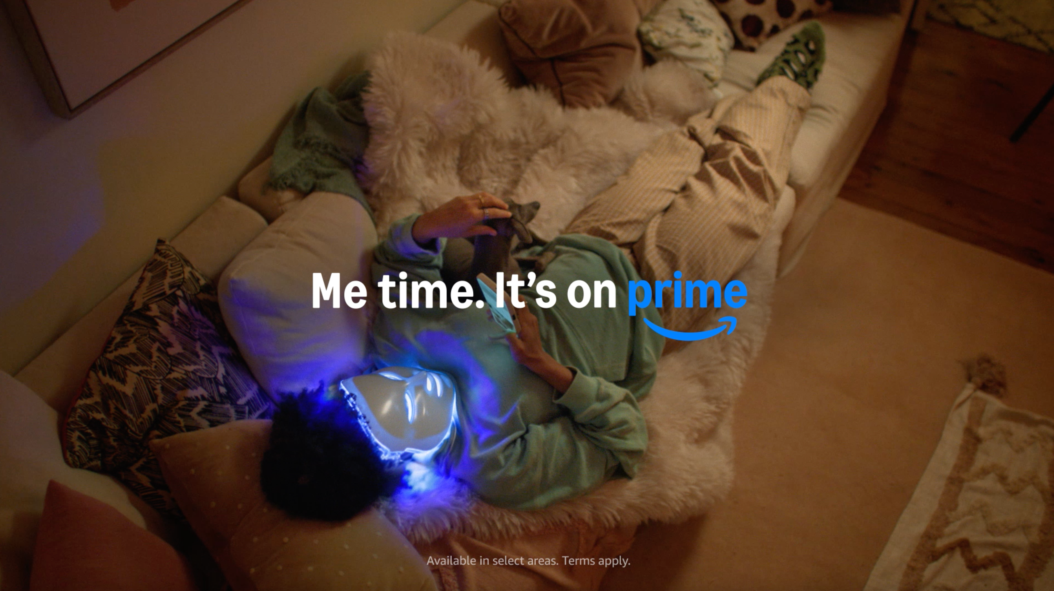A woman in her living room opens an Amazon box containing an LED face mask, text reads "Me Time. Same Day Delivery. It's on Prime"
