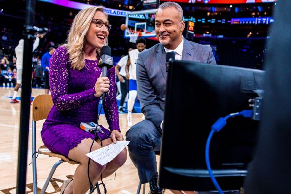 Sixers play-by-play announcer Kate Scott sits on a chair in front of a camera with a colleague.