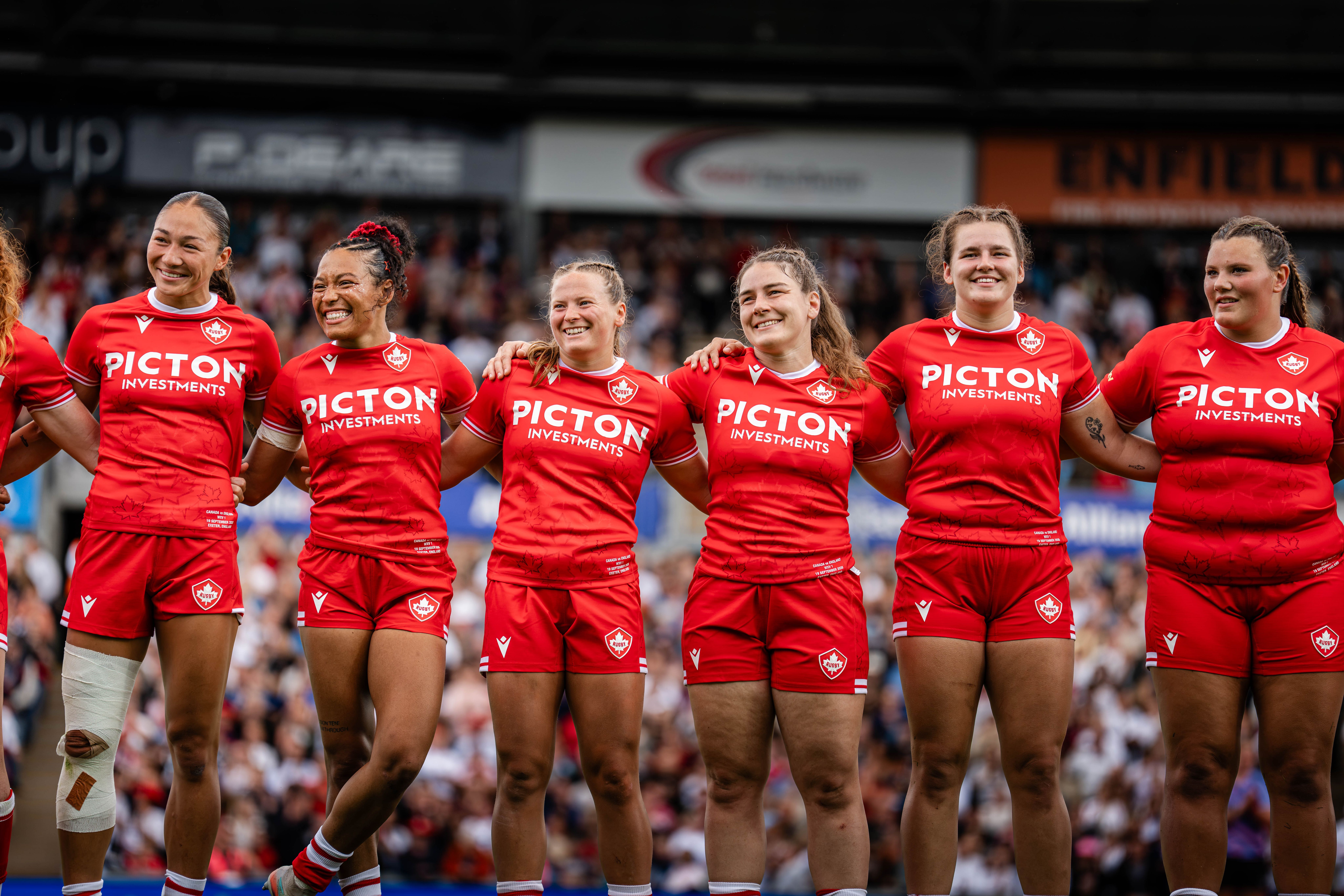 Canadian women’s rugby team standing arm in arm on the field in red jerseys, smiling in front of a stadium crowd.