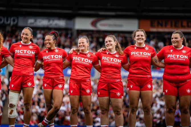 Canadian women’s rugby team standing arm in arm on the field in red jerseys, smiling in front of a stadium crowd.