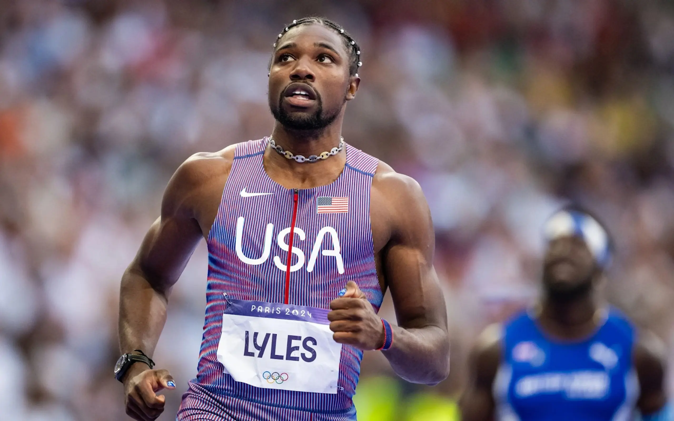 Noah Lyles sports his Team USA uniform, a choker necklace, nail polish, and his signature hair pearls