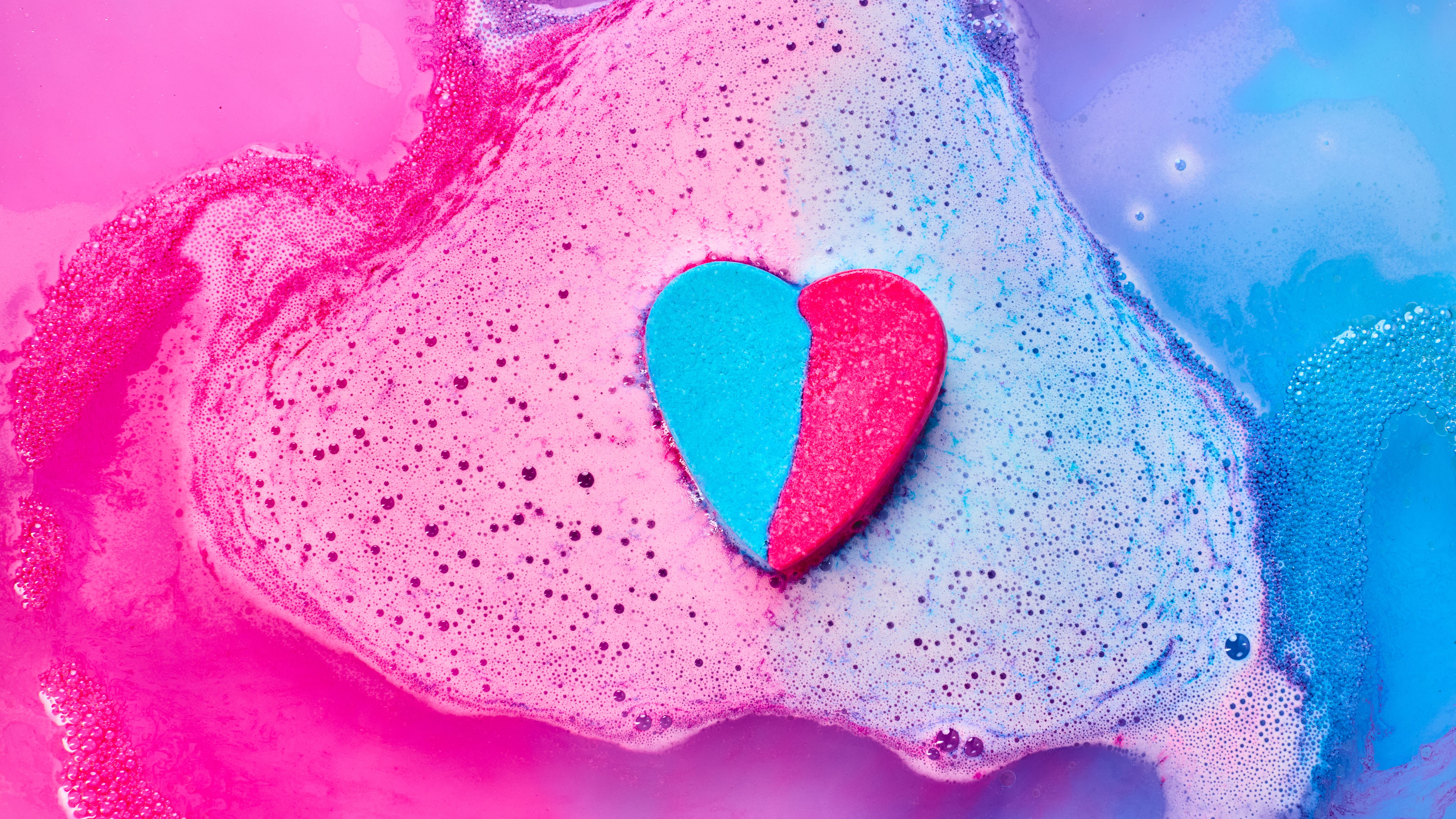 A heart-shaped bath bomb fizzes in water, with pink and blue bubbles.