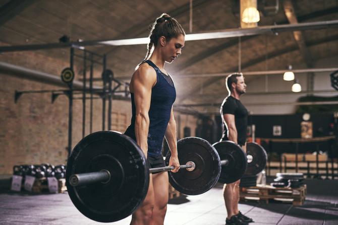 A woman stands in the foreground while a man stands in the background. Both athletes are holding a barbell and preparing to complete a lift.