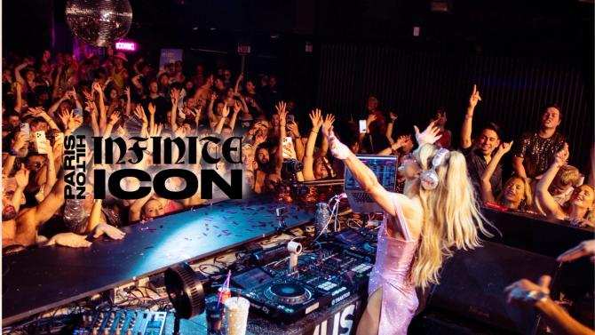 Paris Hilton stands in a DJ booth wearing headphones with her arms outstretched while fans cheer.