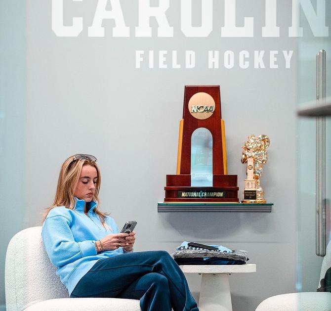UNC field hockey head coach Erin Matson checks her phone while sitting next to one of the team’s national championship trophies.