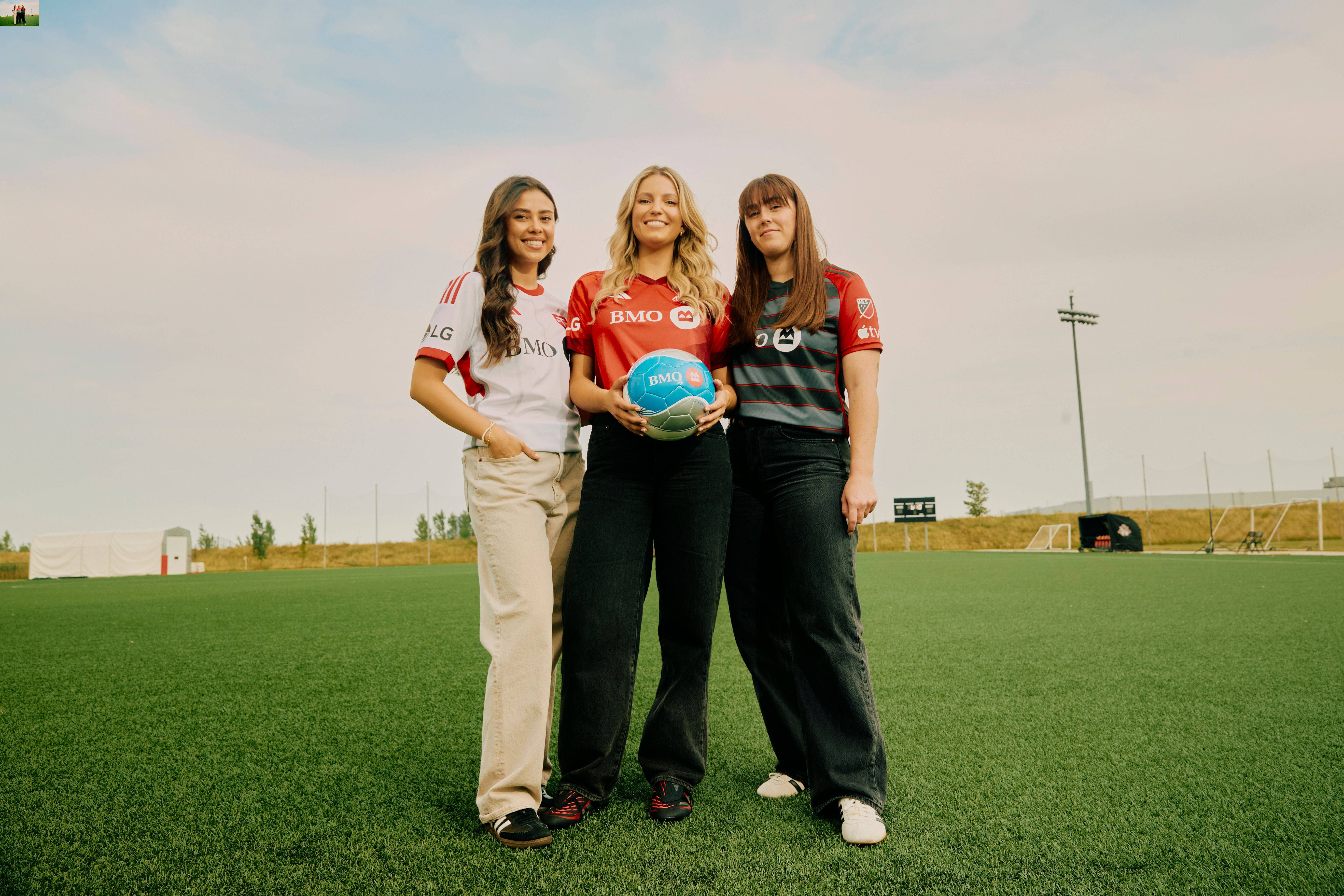 Three women standing on a soccer field, wearing BMO-sponsored soccer jerseys, with one holding a soccer ball.