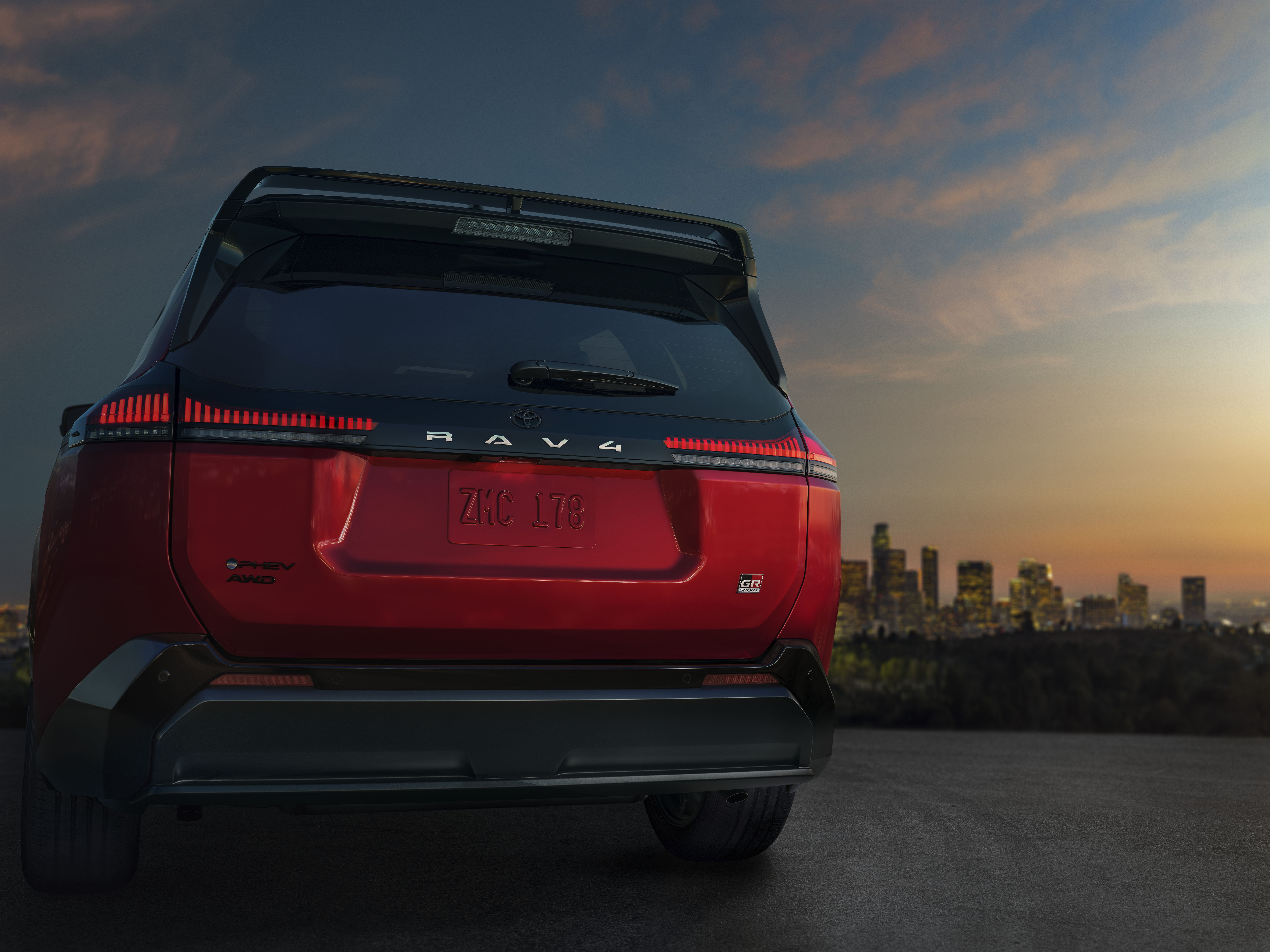 The back of a red 2026 Toyota RAV4 with glowing tail lights sits on top of a gill at dusk with a cityscape in the background.
