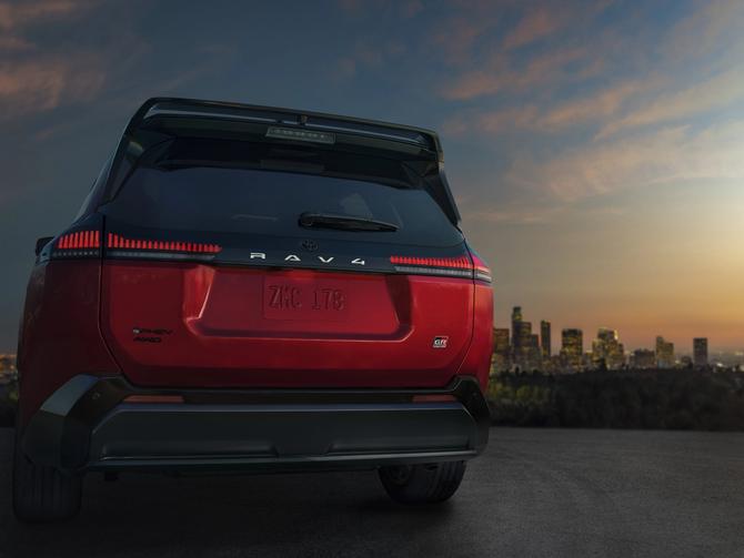 The back of a red 2026 Toyota RAV4 with glowing tail lights sits on top of a gill at dusk with a cityscape in the background.