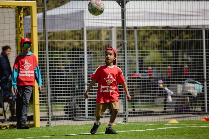 A young boy in a red Jumpstart jersey and backwards cap looks up at a soccer ball in mid-air on an outdoor artificial turf field.