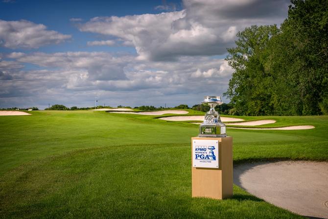 The KPMG Women’s PGA Championship trophy displayed on a pedestal beside a manicured golf course, with rolling fairways, sand bunkers, and a cloudy sky in the background.