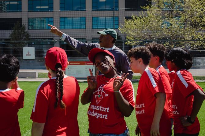 Group of children in red Jumpstart t-shirts gather on a soccer field while a coach points across the field. One child smiles at the camera and flashes peace signs.