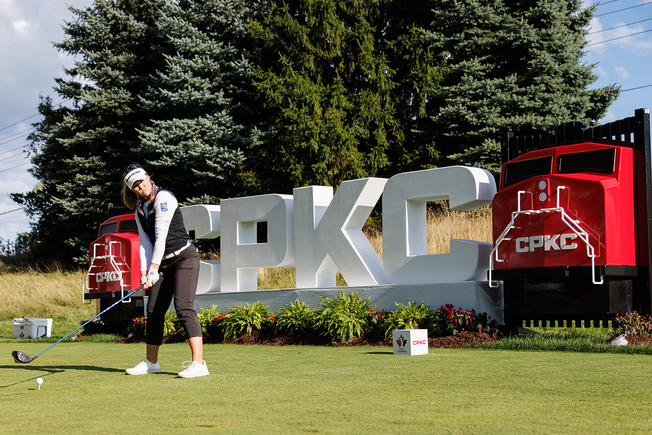 Golfer tees off in front of a large CPKC sign flanked by two red locomotive displays at a golf course.