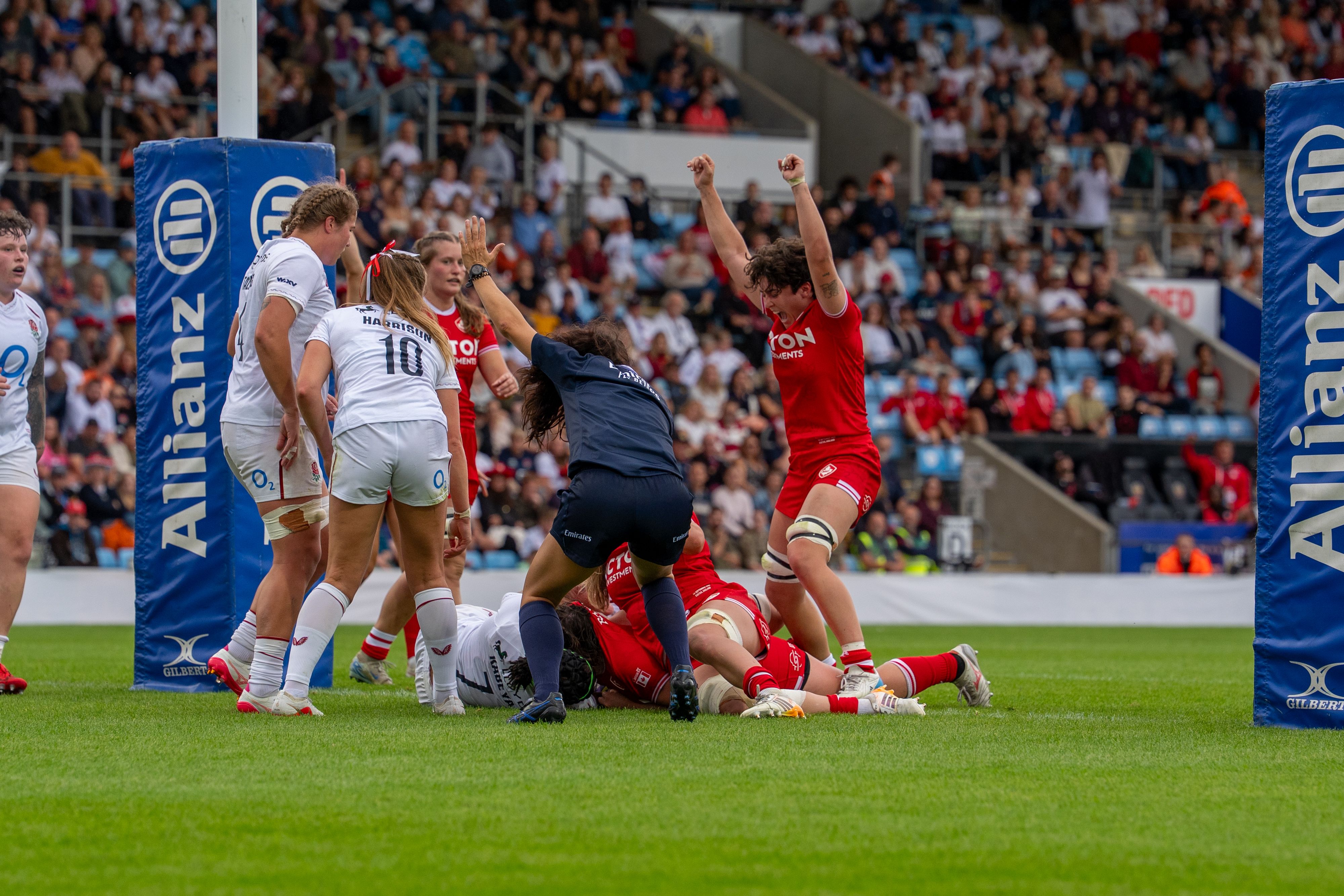 Women’s rugby match in a stadium, with players in red and white jerseys competing near the goal line.