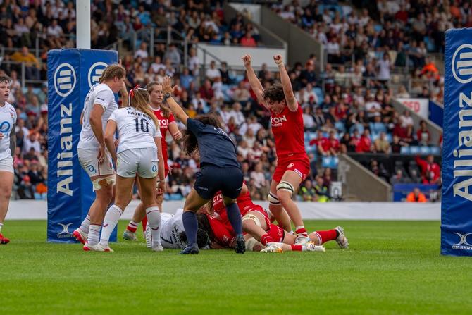 Women’s rugby match in a stadium, with players in red and white jerseys competing near the goal line.