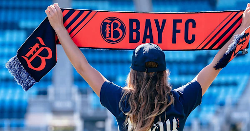 A Bay FC fan holds up a scarf with the team’s branding displayed across it.