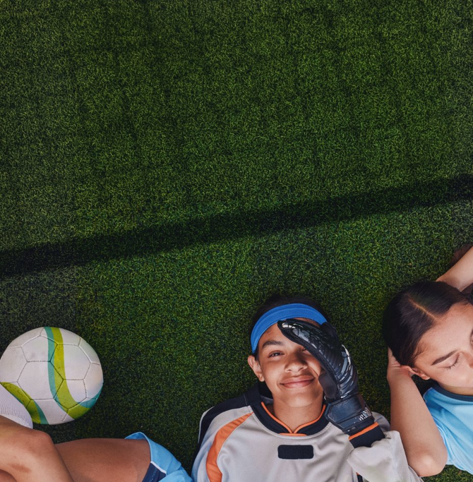 Three young soccer players lie on a grassy field beside a soccer ball, with one smiling at the camera while wearing goalkeeper gloves.