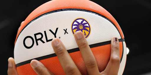A manicured hand holds a WNBA basketball emblazoned with the logos of Orly and the Los Angeles Sparks.