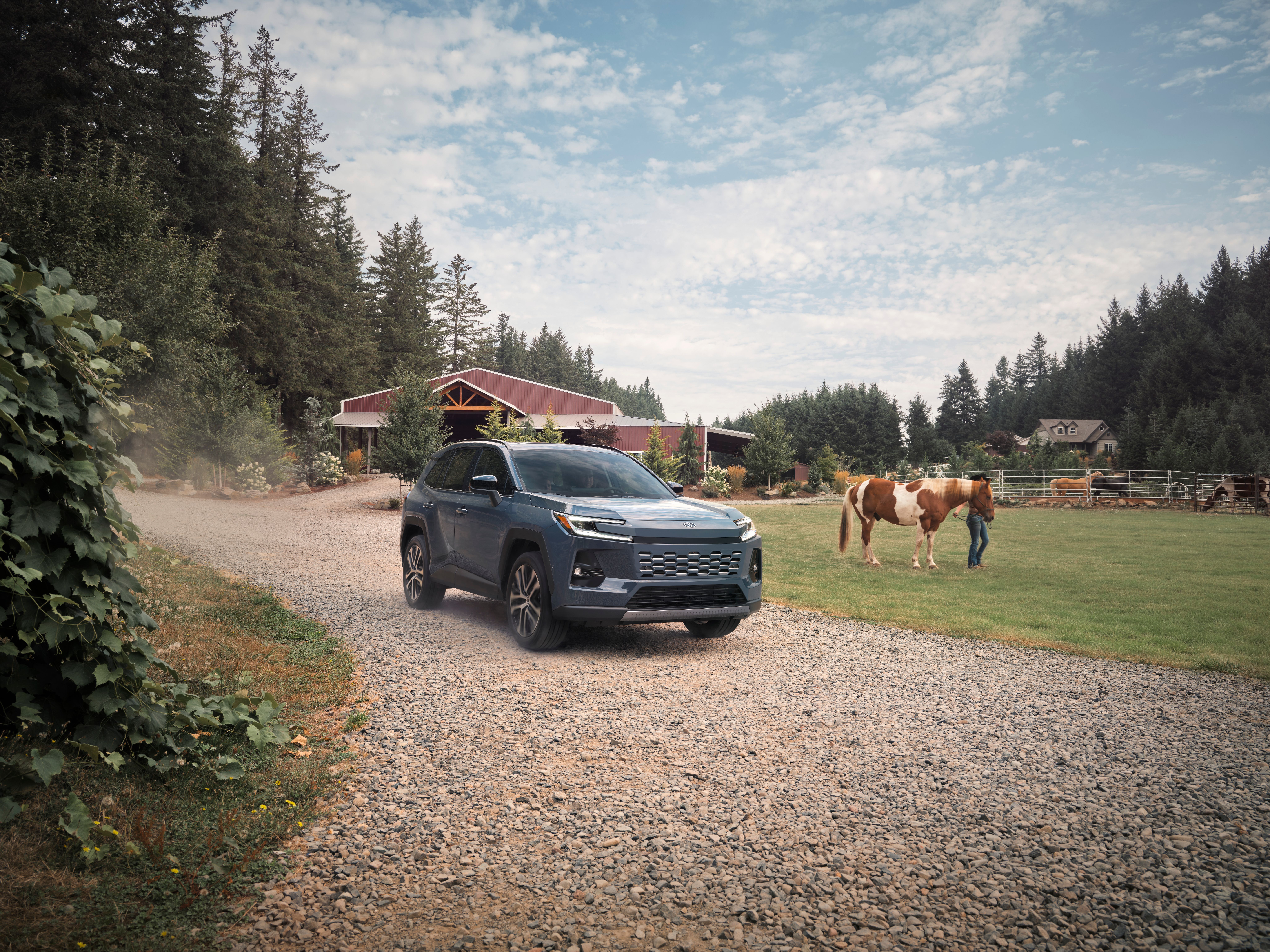 A blue Toyota RAV4 Limited drives along a gravel road through a rural property, with a barn, horses, and tree-lined fields creating a peaceful countryside backdrop.
