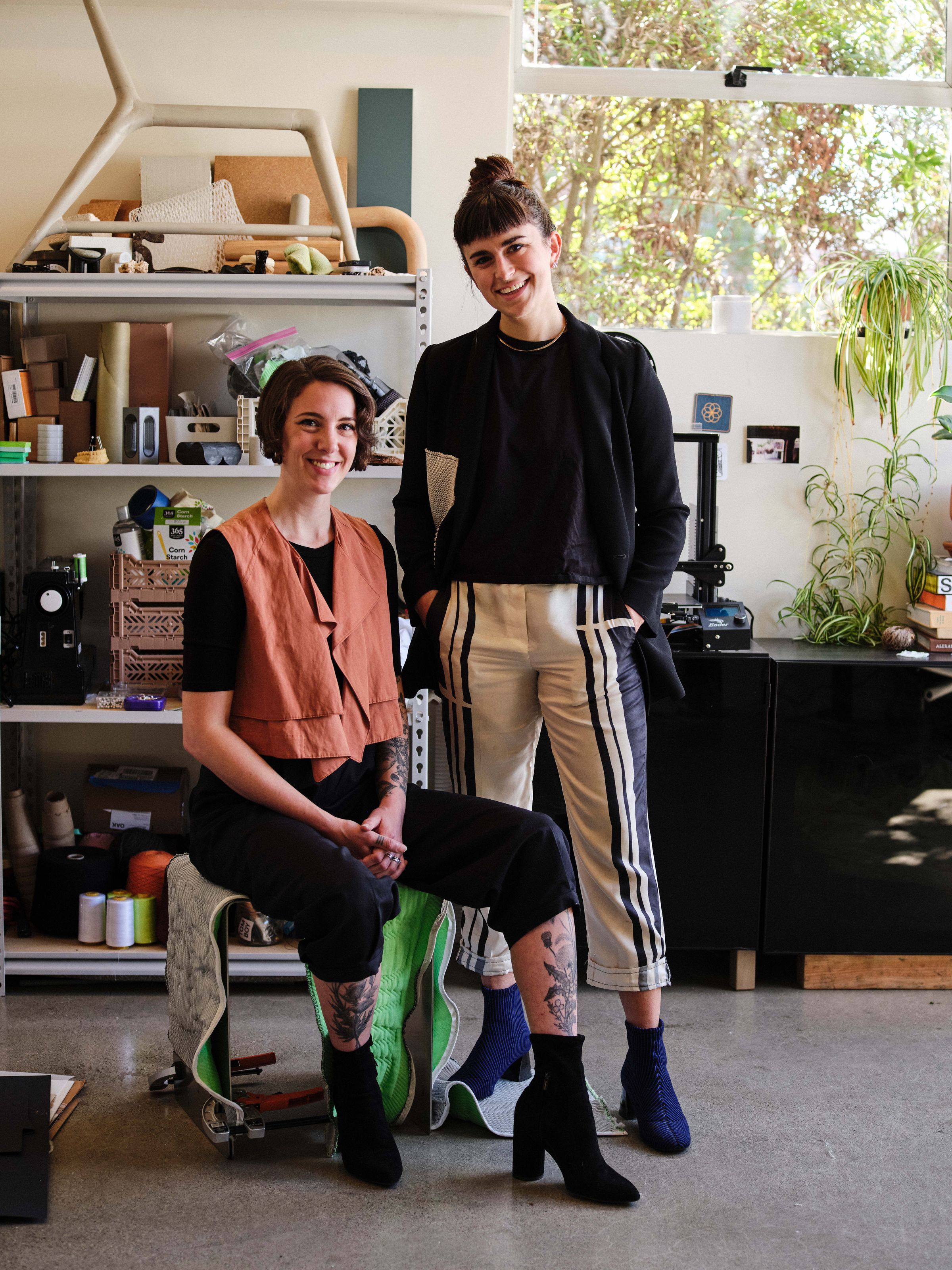 Baillie sitting on a stool and Lauryn standing behind looking at the camera. They are standing in a studio in front of shelves that are covered in materials and ingredients. 