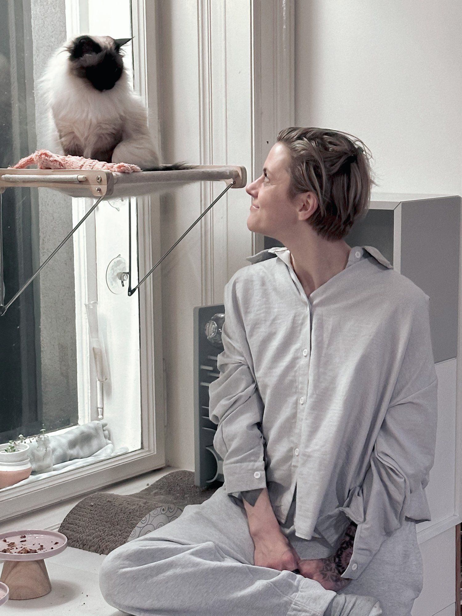 Claudia Six sitting cross-legged at a window, looking at a fluffy cat perched above her — quiet mutual curiosity