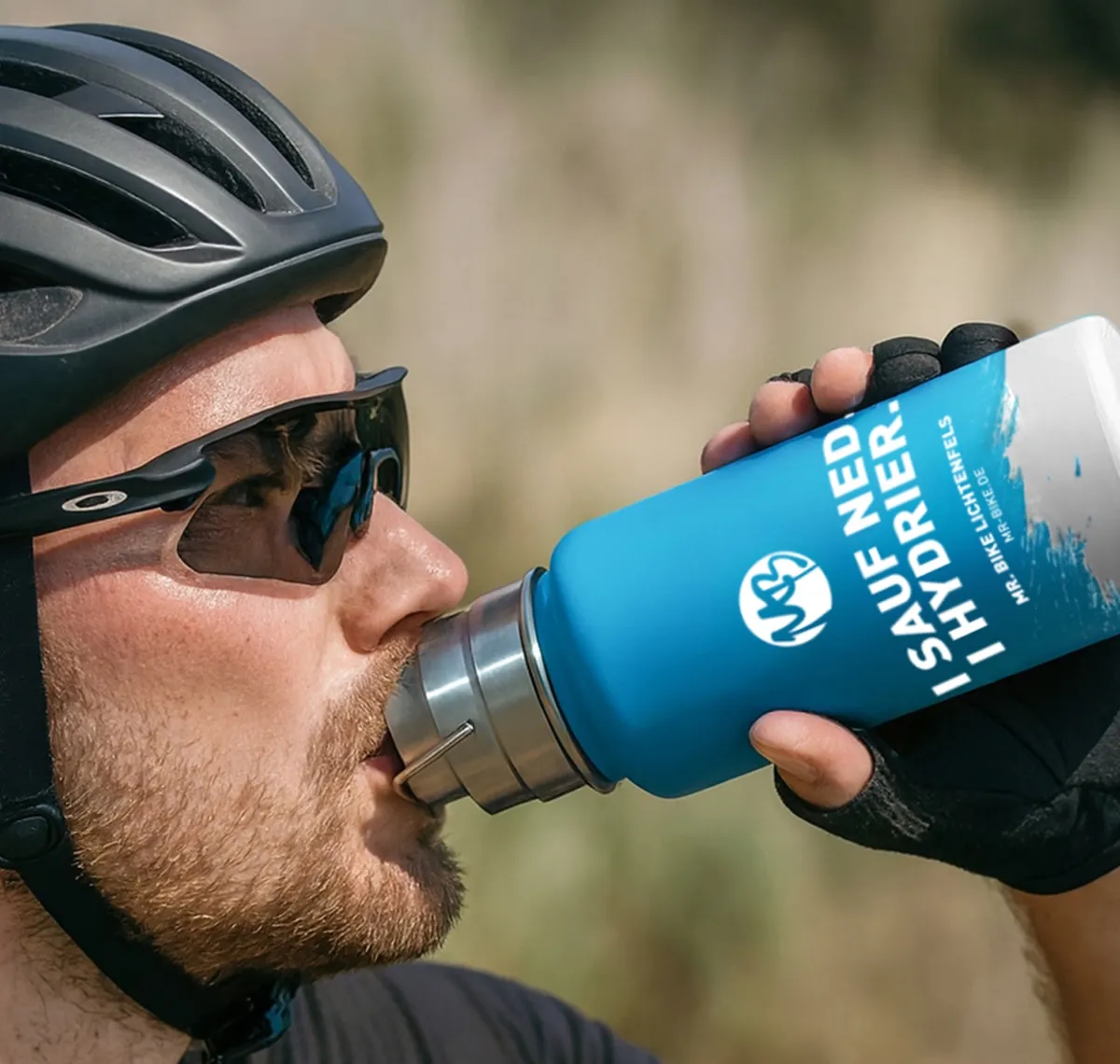 Mr. Bike: Radfahrer mit Helm und Sonnenbrille trinkt aus einer blau bedruckten Trinkflasche mit dem Aufdruck „Sauf ned, hydrier"