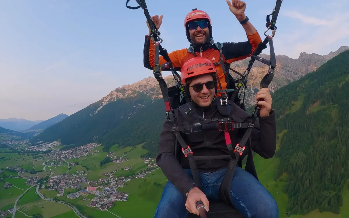 Fly Stubai: Pilot und Gast hängen im Tandem-Gurtzeug hoch über dem grünen Stubaital, beide mit roten Helmen, dahinter die Berge im Abendlicht.