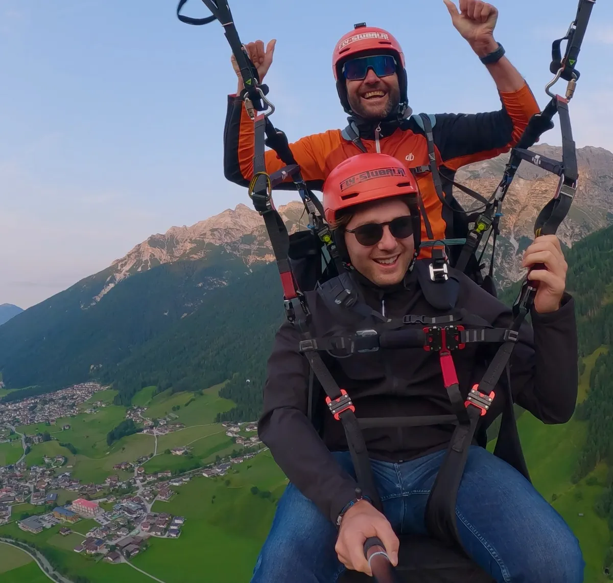 Fly Stubai: Pilot und Gast hängen im Tandem-Gurtzeug hoch über dem grünen Stubaital, beide mit roten Helmen, dahinter die Berge im Abendlicht.