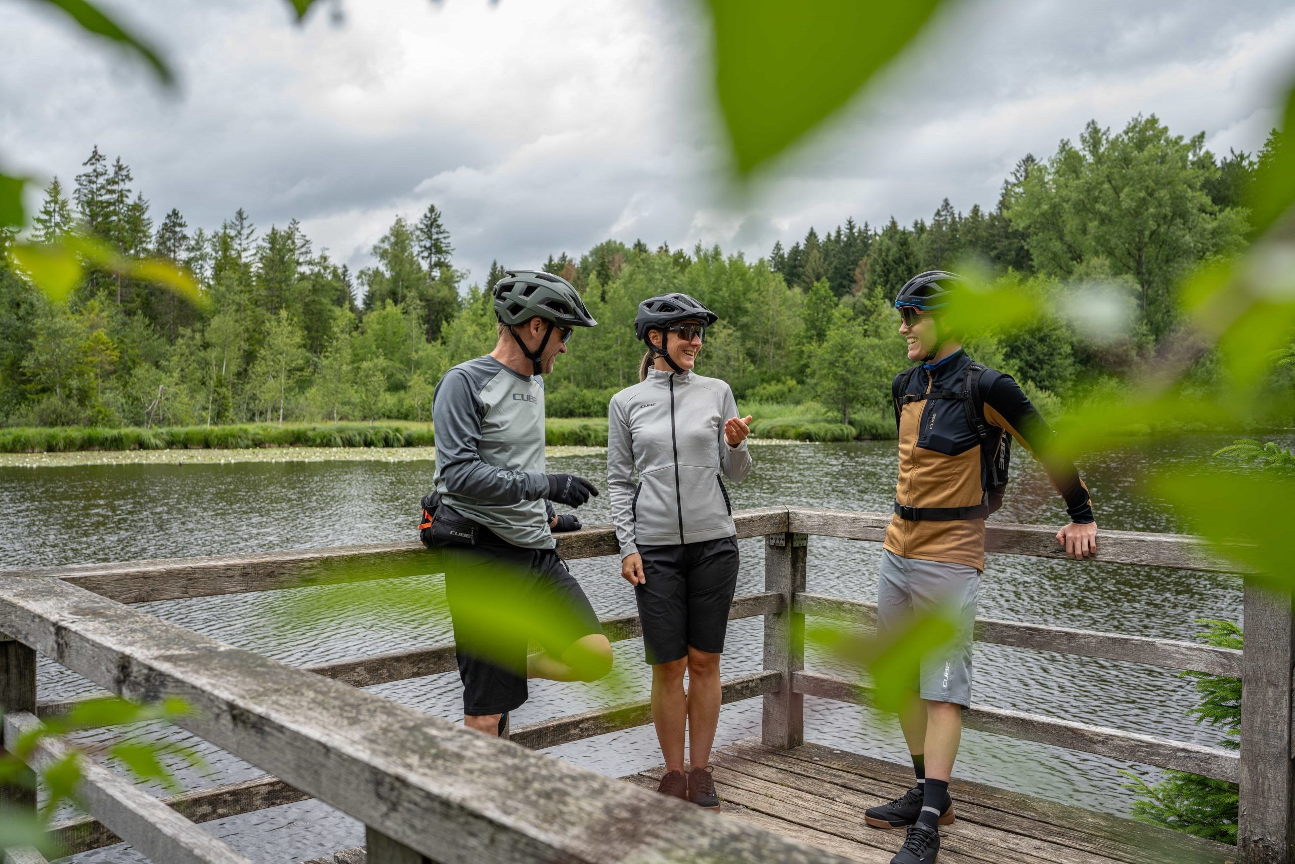 Employer Branding für den CUBE Store Lindenberg: drei Radfahrerinnen und Radfahrer stehen auf einem Holzsteg am See und unterhalten sich.