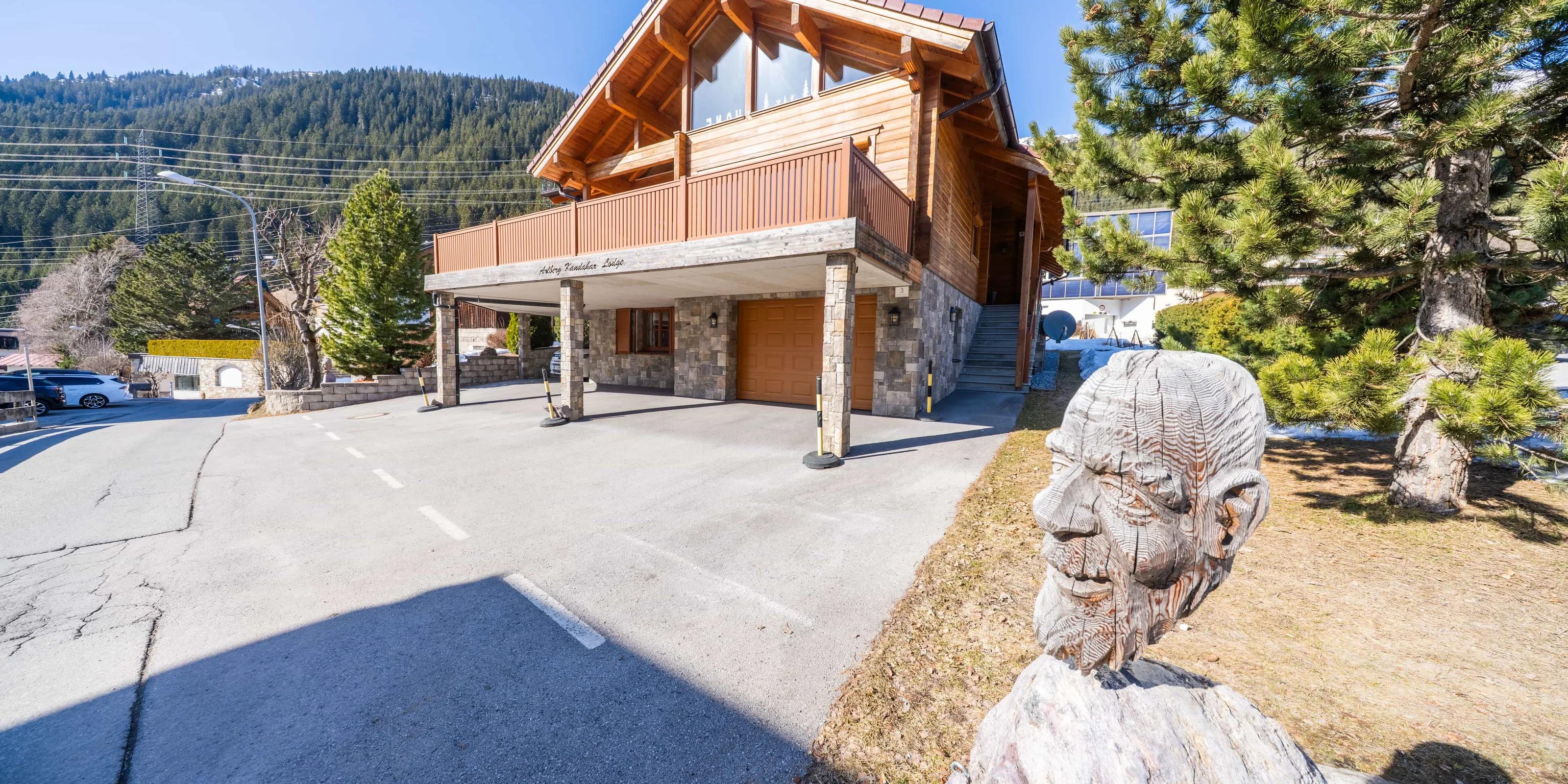 Fotoproduktion: Die Arlberg Kandahar Lodge von der Straße aus: das Holzhaus mit überdachtem Vorplatz, davor eine geschnitzte Holzfigur, dahinter der bewaldete Hang