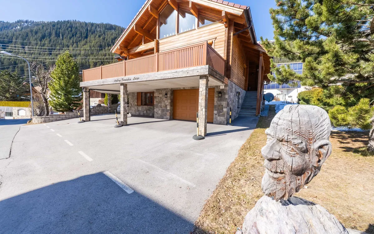 Fotoproduktion: Die Arlberg Kandahar Lodge von der Straße aus: das Holzhaus mit überdachtem Vorplatz, davor eine geschnitzte Holzfigur, dahinter der bewaldete Hang