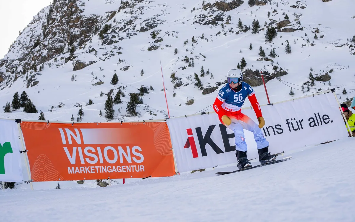 Crazy Curves / FIS Para Snowboard Worldcup: Para-Snowboarder mit Startnummer 56 auf der Rennpiste, davor die orange Bande der Marketingagentur Invisions