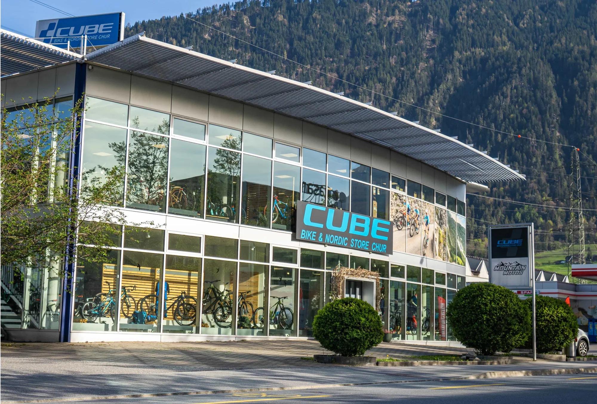CUBE STORE CHUR: Der CUBE Bike & Nordic Store Chur von außen, Glasfassade mit Bikes im Schaufenster, dahinter bewaldete Berghänge