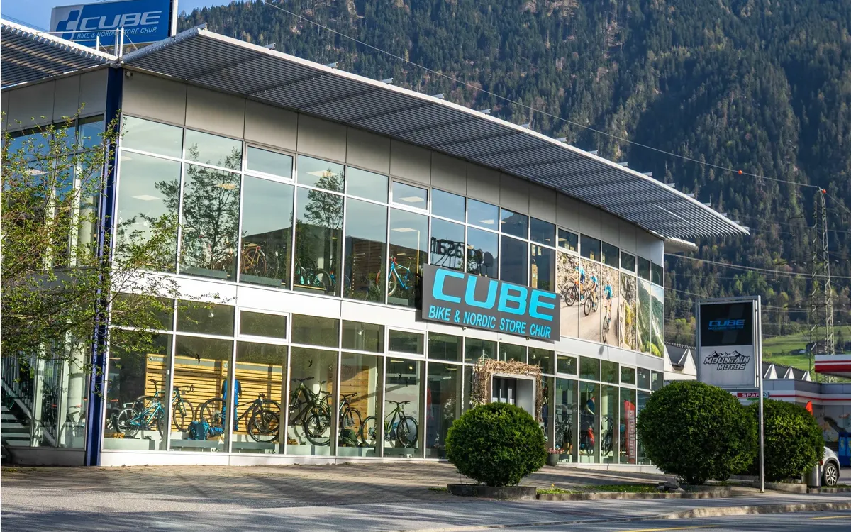 CUBE STORE CHUR: Der CUBE Bike & Nordic Store Chur von außen, Glasfassade mit Bikes im Schaufenster, dahinter bewaldete Berghänge