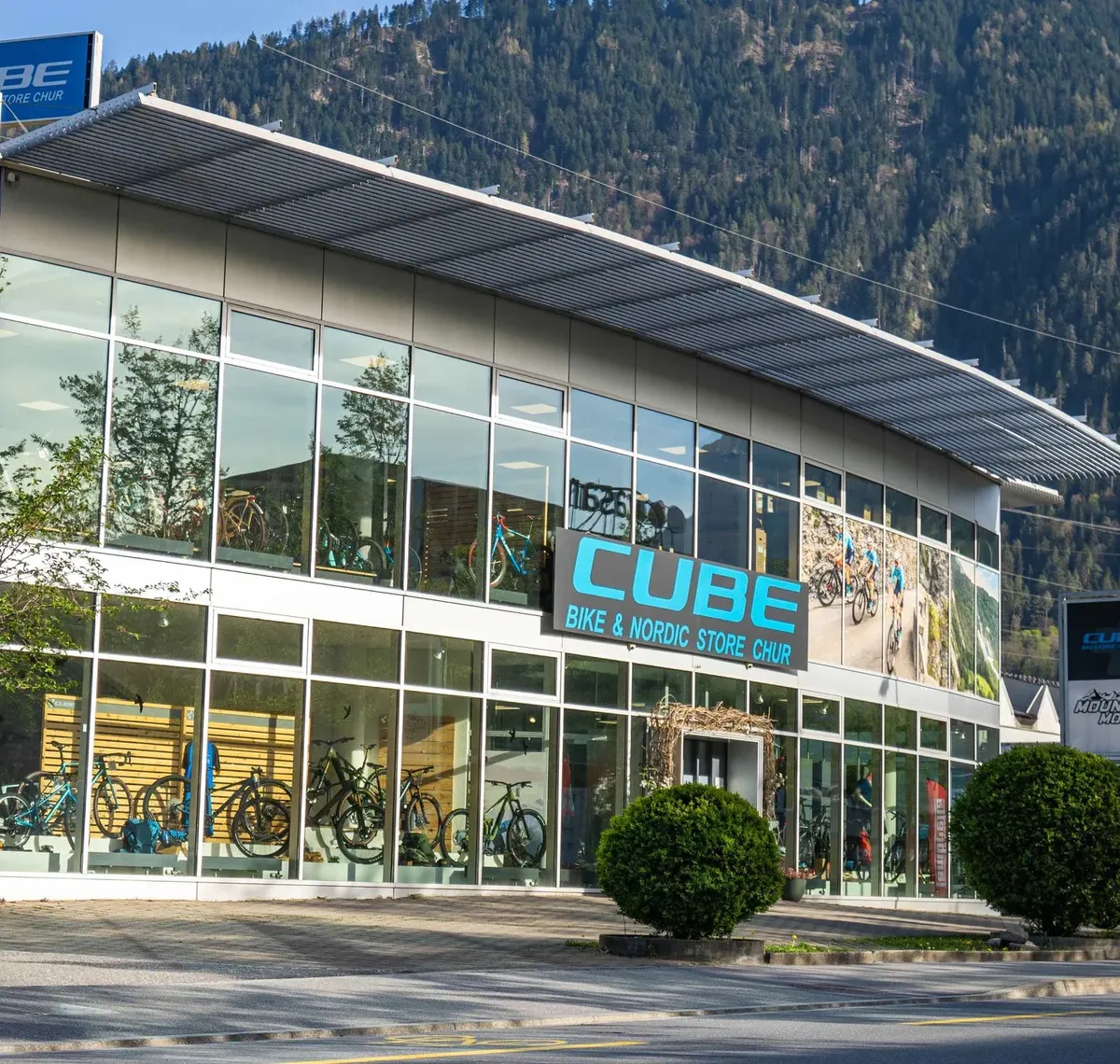 CUBE STORE CHUR: Der CUBE Bike & Nordic Store Chur von außen, Glasfassade mit Bikes im Schaufenster, dahinter bewaldete Berghänge