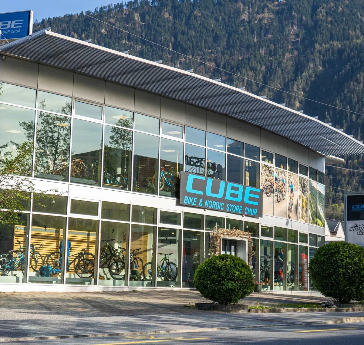 CUBE STORE CHUR: Der CUBE Bike & Nordic Store Chur von außen, Glasfassade mit Bikes im Schaufenster, dahinter bewaldete Berghänge