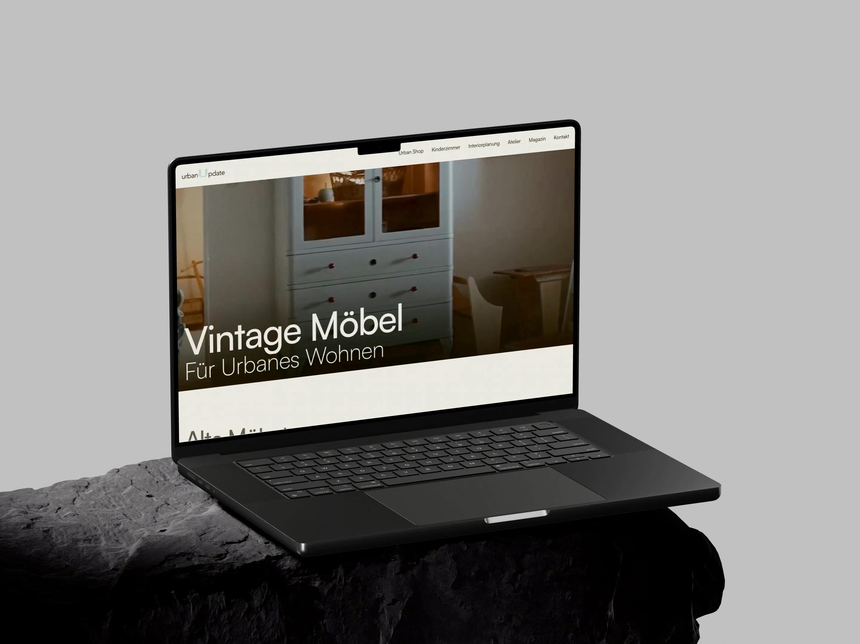 UrbanUpdate: Ein aufgeklapptes MacBook Pro auf einem dunklen Steinsockel vor hellgrauer Wand zeigt die Startseite von UrbanUpdate mit einem hellblauen Vintage-Kasten und der Zeile Vintage Möbel für urbanes Wohnen.