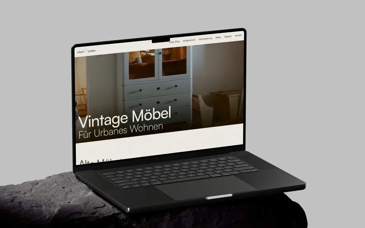 UrbanUpdate: Ein aufgeklapptes MacBook Pro auf einem dunklen Steinsockel vor hellgrauer Wand zeigt die Startseite von UrbanUpdate mit einem hellblauen Vintage-Kasten und der Zeile Vintage Möbel für urbanes Wohnen.