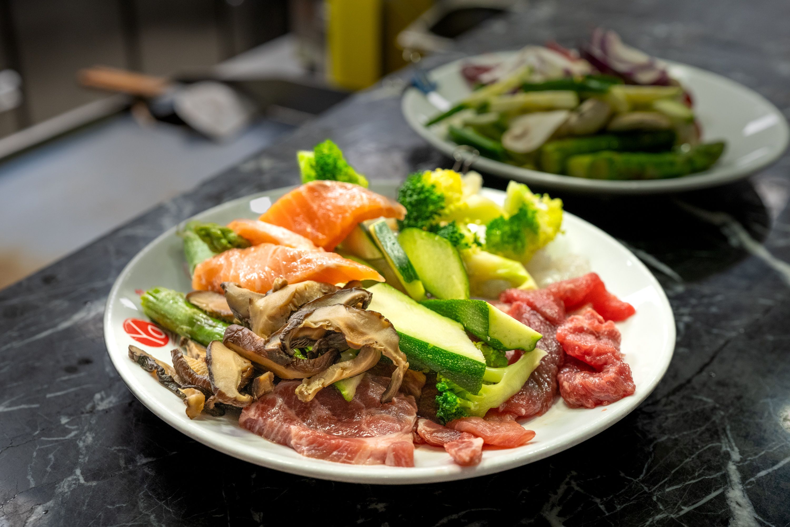 Foodfotografie Buffet und Karte: Ein weisser Teller mit Lachs, Rindfleisch, Pilzen, Zucchini und Brokkoli auf schwarzer Marmorfläche, dahinter angeschnitten ein zweiter Teller.