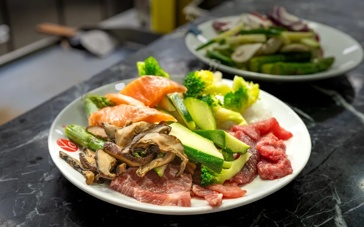 Foodfotografie Buffet und Karte: Ein weisser Teller mit Lachs, Rindfleisch, Pilzen, Zucchini und Brokkoli auf schwarzer Marmorfläche, dahinter angeschnitten ein zweiter Teller.
