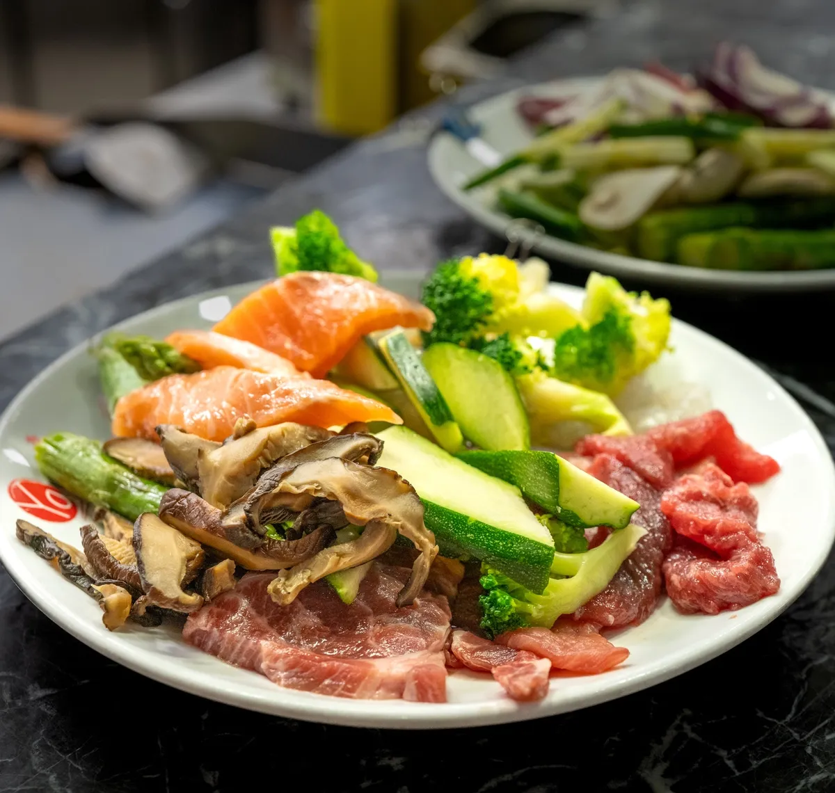 Asia Restaurant YAMI: Ein weisser Teller mit Lachs, Rindfleisch, Pilzen, Zucchini und Brokkoli auf schwarzer Marmorfläche, dahinter angeschnitten ein zweiter Teller.