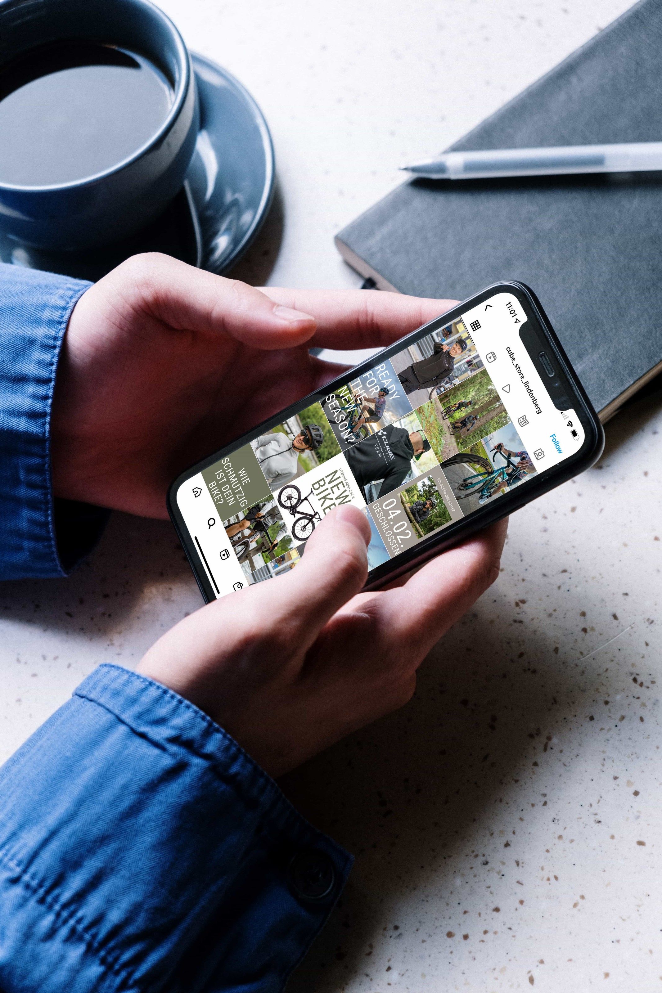 CUBE Store Lindenberg: Zwei Hände halten ein Smartphone mit einem Instagram-Feed über einem Schreibtisch mit Kaffeetasse und Notizbuch