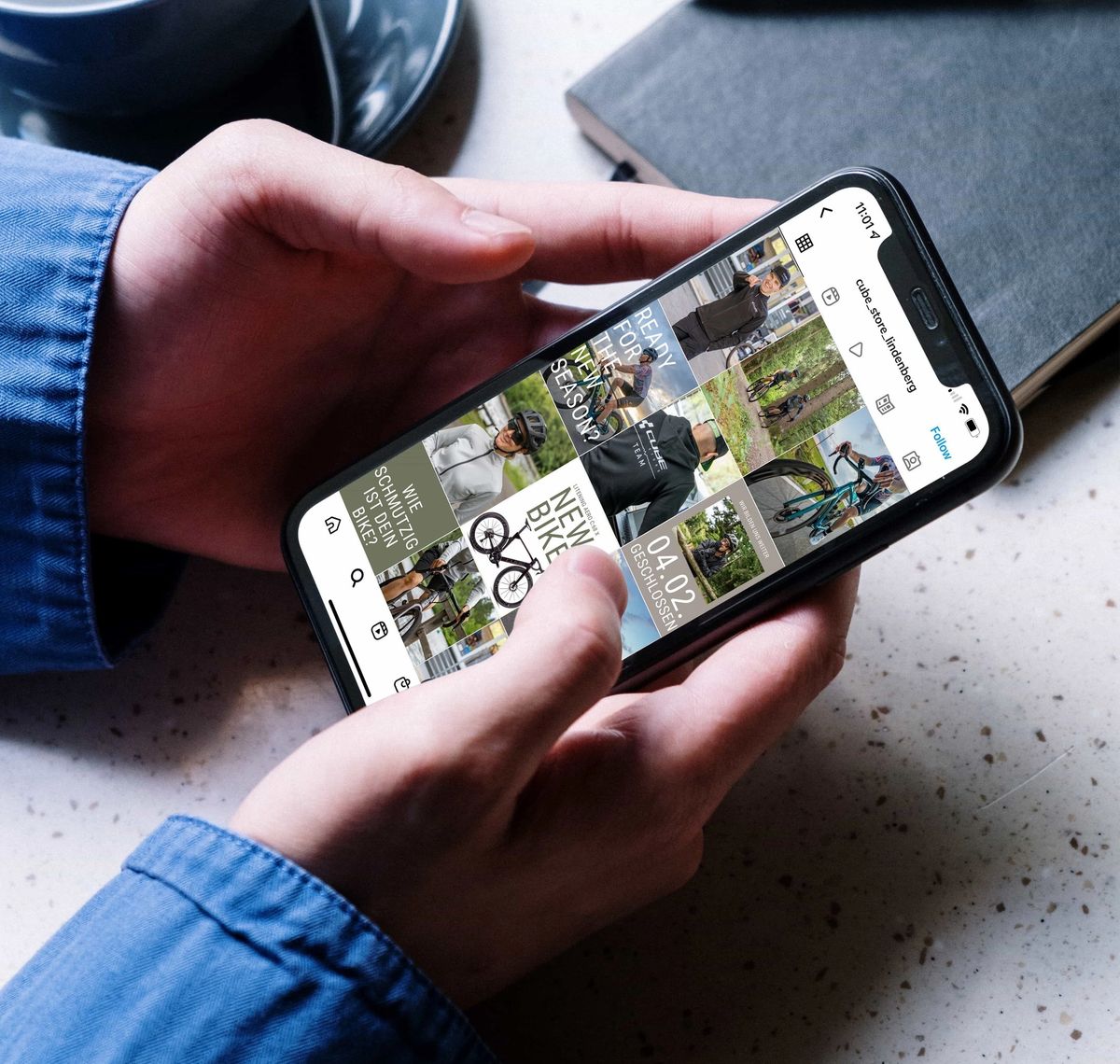 CUBE Store Lindenberg: Zwei Hände halten ein Smartphone mit einem Instagram-Feed über einem Schreibtisch mit Kaffeetasse und Notizbuch