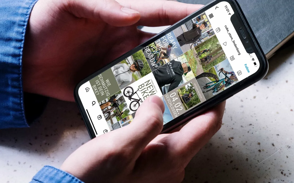 CUBE Store Lindenberg: Zwei Hände halten ein Smartphone mit einem Instagram-Feed über einem Schreibtisch mit Kaffeetasse und Notizbuch