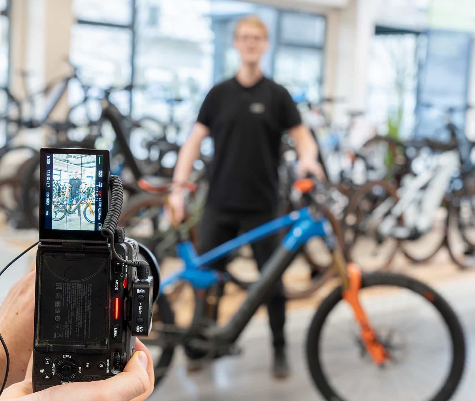 Fachhandelspartner: Blick über das Display einer Filmkamera auf einen Mitarbeiter, der ein Mountainbike durch den Verkaufsraum schiebt