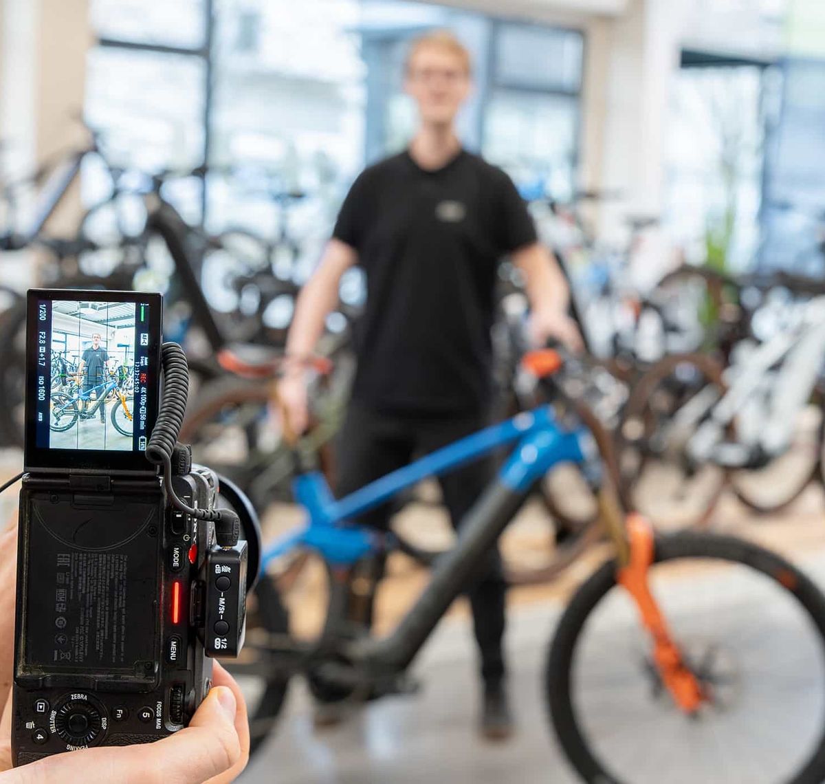 Fachhandelspartner: Blick über das Display einer Filmkamera auf einen Mitarbeiter, der ein Mountainbike durch den Verkaufsraum schiebt
