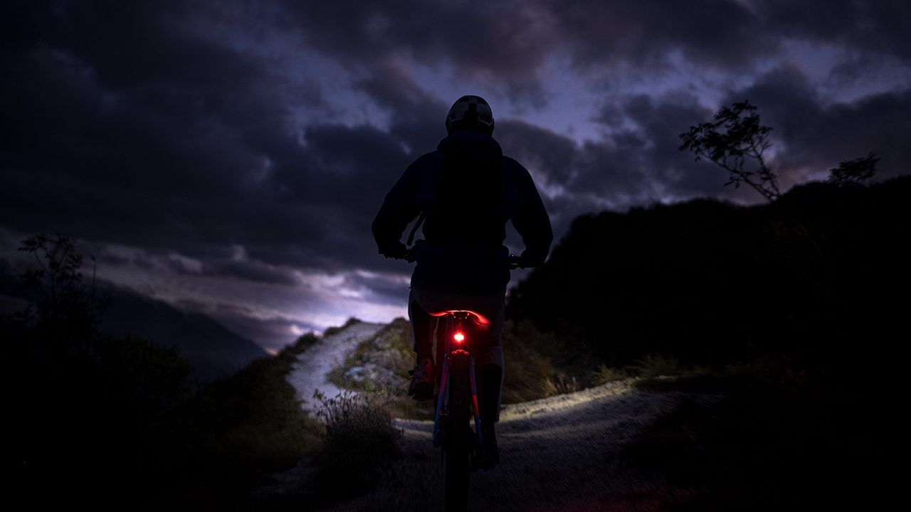 Litemove: Mountainbiker bei Dämmerung auf einem Schotterweg, das rote Rücklicht leuchtet, darüber ein Gewitterhimmel