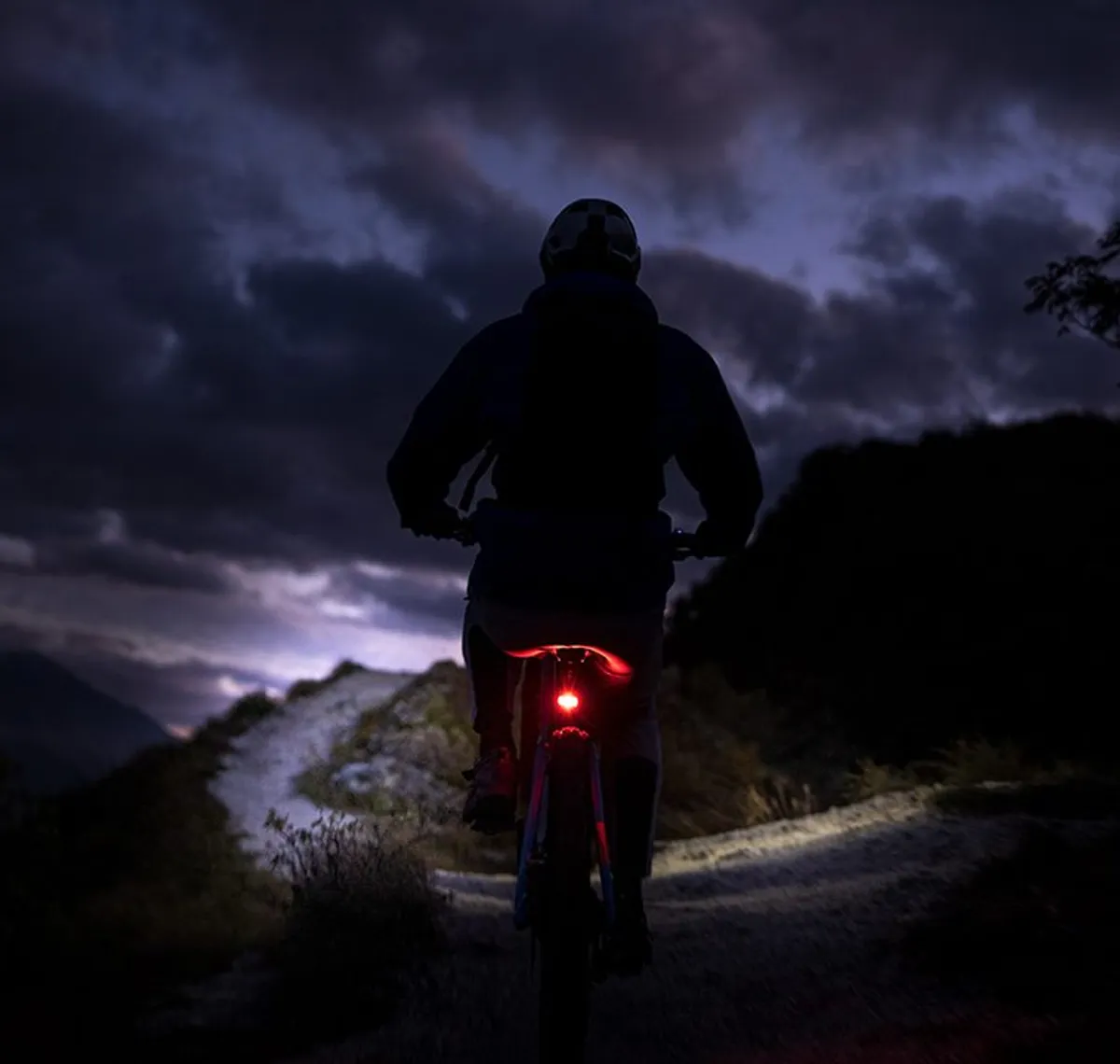 Litemove: Mountainbiker bei Dämmerung auf einem Schotterweg, das rote Rücklicht leuchtet, darüber ein Gewitterhimmel