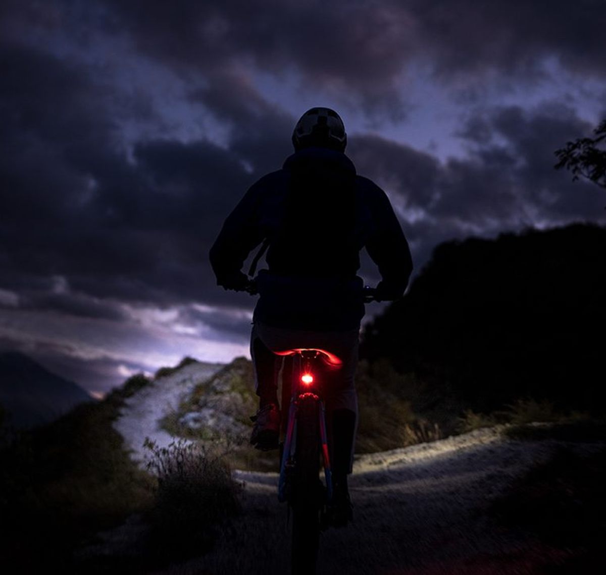 Litemove: Mountainbiker bei Dämmerung auf einem Schotterweg, das rote Rücklicht leuchtet, darüber ein Gewitterhimmel
