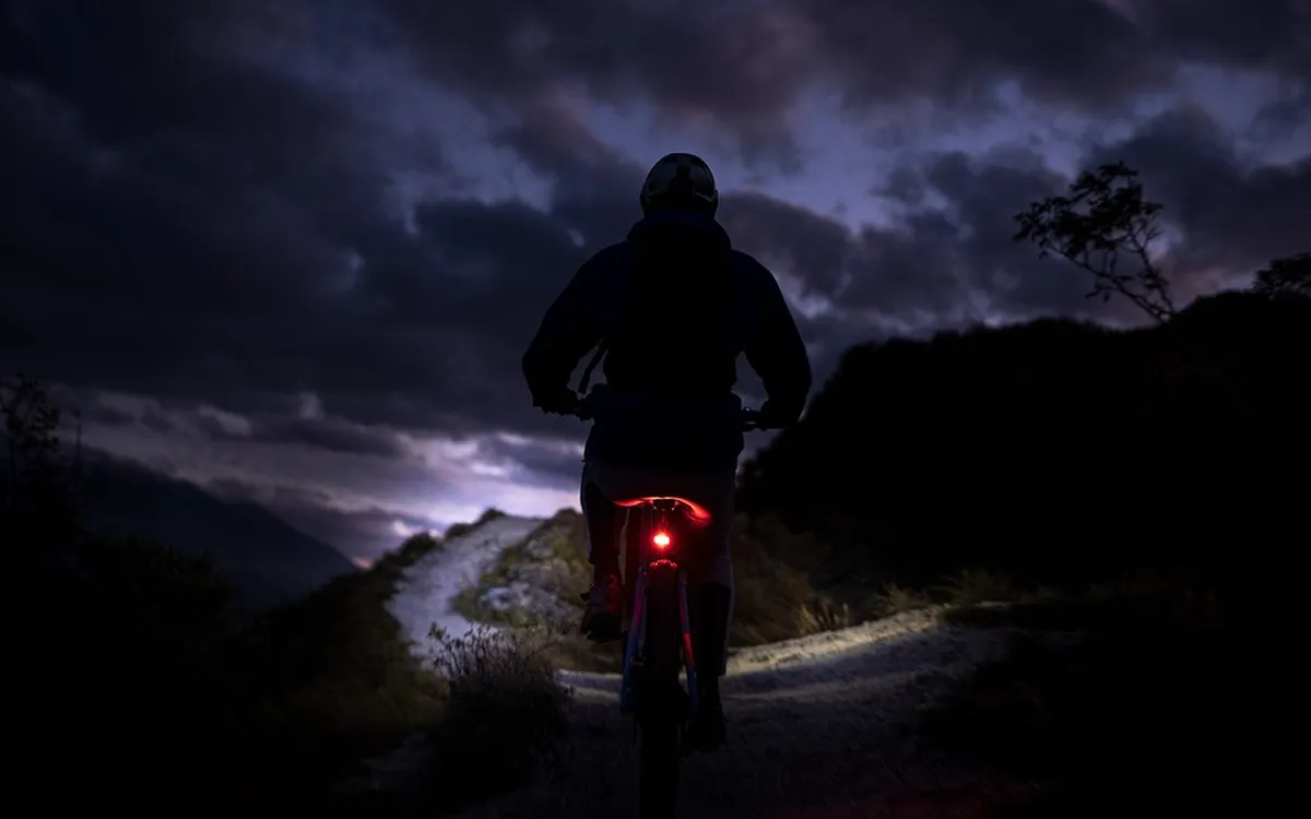 Litemove: Mountainbiker bei Dämmerung auf einem Schotterweg, das rote Rücklicht leuchtet, darüber ein Gewitterhimmel