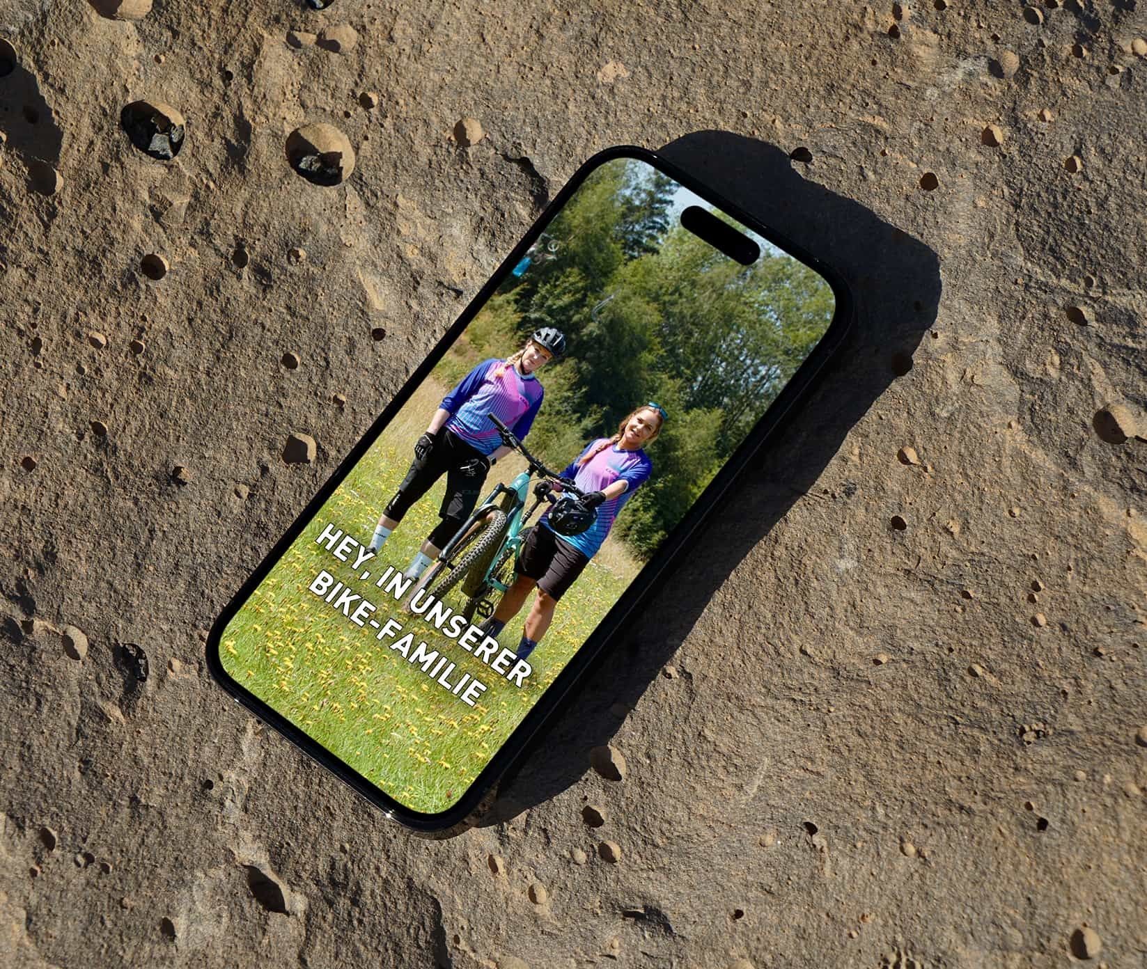 V&S Bike GmbH: Smartphone auf einem Felsen zeigt einen Social-Media-Beitrag mit zwei Radfahrerinnen und der Zeile „Hey in unserer Bike-Familie"